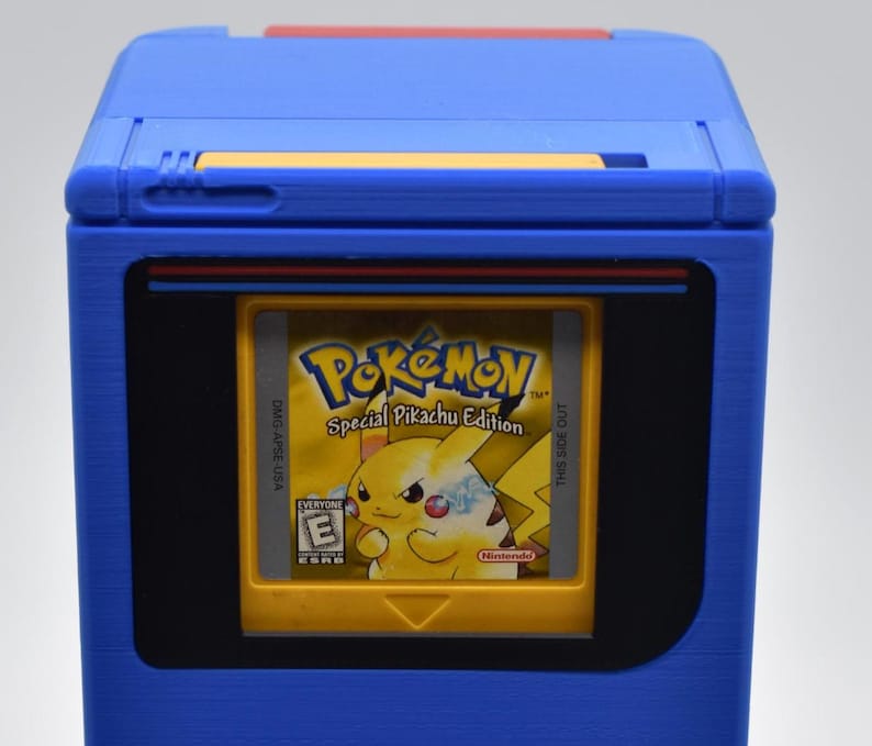 以下が含まれることがあります： 青いゲームボーイカートリッジホルダーと黄色のポケモンSpecial Pikachu Editionカートリッジ。ホルダーは長方形のデザインで、黒い画面領域と上部に赤いアクセントがあります。カートリッジは黄色で、ピカチュウのグラフィックが付いています。