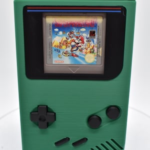 以下が含まれることがあります： スーパーマリオブラザーズのゲームカートリッジが挿入された緑色の携帯ゲーム機。コンソールには方向パッド、2つのアクションボタン、ゲームのグラフィックを表示する画面があります。レトロゲームアイテムです。