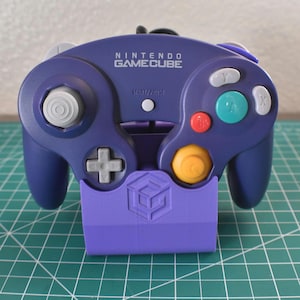 Könnte beinhalten: Ein violetter Nintendo GameCube-Controller mit passendem lila Ständer. Der Controller hat verschiedenfarbige Tasten, darunter rot, grün und gelb. Der Text "NINTENDO GAMECUBE" ist auf dem Controller zu sehen.