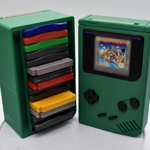 以下が含まれることがあります： 緑色のレトロスタイルのゲーム機ケースとカートリッジホルダー。ケースは、ゲームを表示する画面を備えた、クラシックなハンドヘルドコンソールに似ています。カートリッジホルダーには、カラフルなゲームカートリッジが詰まっています。