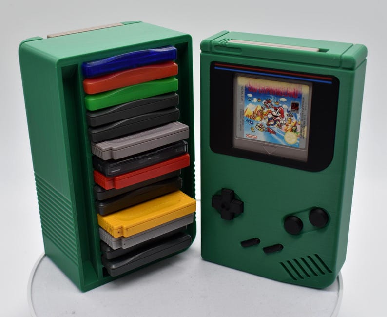 以下が含まれることがあります： 緑色のレトロスタイルのゲーム機ケースとカートリッジホルダー。ケースは、ゲームを表示する画面を備えた、クラシックなハンドヘルドコンソールに似ています。カートリッジホルダーには、カラフルなゲームカートリッジが詰まっています。