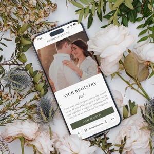 May include: A smartphone displaying a wedding registry website with the text Sophia & Jacob and Our Registry. The screen shows a photo of a couple and a button labelled View Registry. The phone is surrounded by white roses and green foliage.