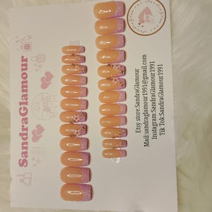 Puede incluir: Un conjunto de 10 uñas postizas con una base rosa claro y una punta lavanda. Cada uña tiene un pequeño grupo de purpurina dorada y un pequeño diseño de flor.