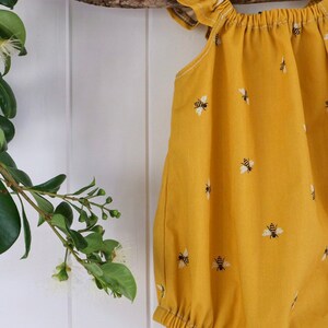 May include: A yellow baby onesie with a bee pattern. The onesie has ruffles on the shoulders and a gathered neckline.