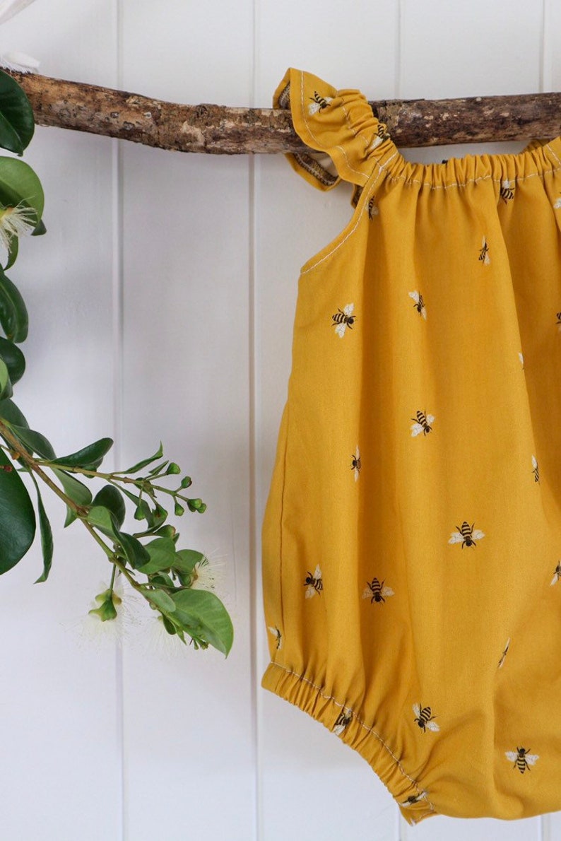 May include: A yellow baby onesie with a bee pattern. The onesie has ruffles on the shoulders and a gathered neckline.