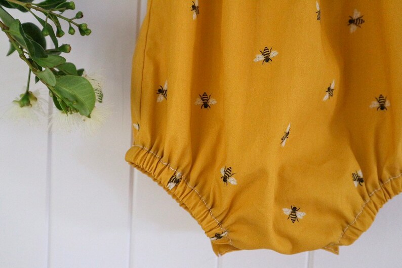 May include: A yellow baby onesie with a bee pattern. The onesie has a gathered elastic waistband.