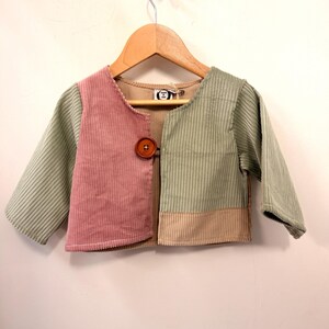 May include: A colour-blocked corduroy jacket featuring a dusty pink left panel, a sage green right panel, and a tan lower section. The jacket has long sleeves, a round neckline, and a single large brown button closure. A label reads Stitched by Love.
