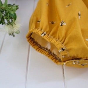 May include: Close-up of the bottom of a yellow baby onesie with a bee print. The onesie has a gathered elastic waistband and snap closures.