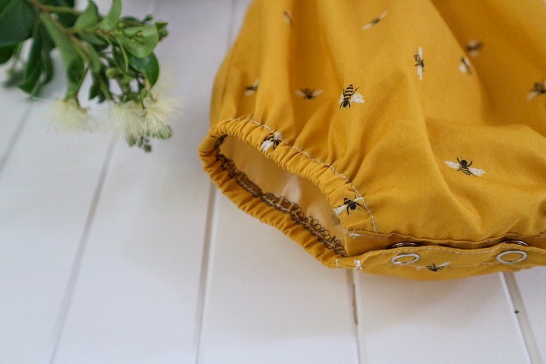 May include: Close-up of the bottom of a yellow baby onesie with a bee print. The onesie has a gathered elastic waistband and snap closures.