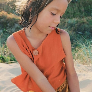 May include: A young person wearing a bright orange sleeveless top with a single brown button and matching mustard yellow trousers. The person is sitting on a sandy surface with green grass in the background.