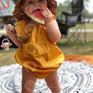 May include: A baby girl wearing a yellow dress with a bee pattern and a straw hat is eating a slice of watermelon. She is standing on a white and black patterned blanket.