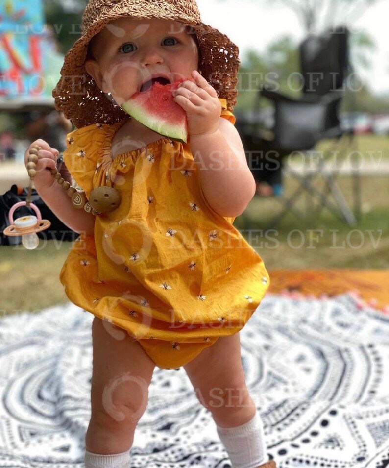 May include: A baby girl wearing a yellow dress with a bee pattern and a straw hat is eating a slice of watermelon. She is standing on a white and black patterned blanket.