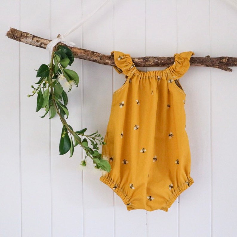 May include: A yellow baby onesie with a bee pattern. The onesie has ruffles on the shoulders and a gathered skirt.