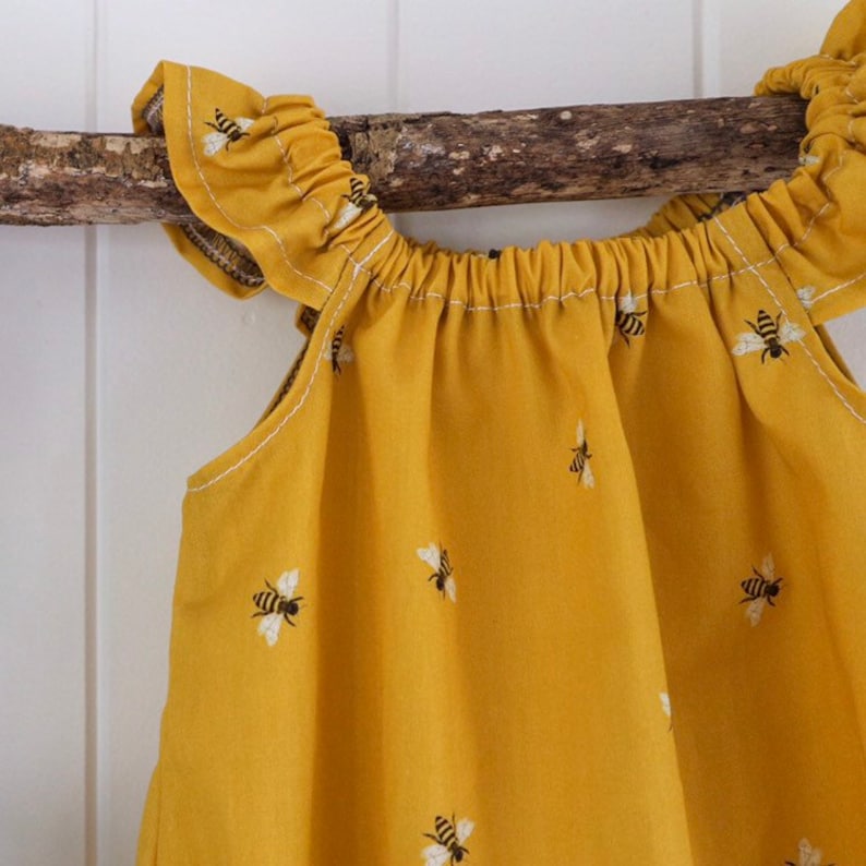 May include: A yellow sleeveless dress with a ruffled neckline and a pattern of small black and white bees. The dress is hanging on a branch.