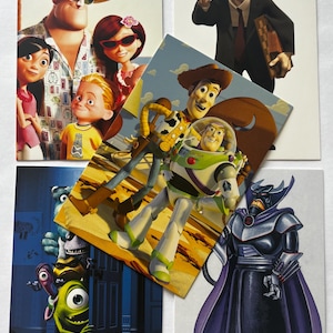 Può includere: Una collezione di cinque cartoline rettangolari con vari personaggi animati. Le immagini includono la famiglia degli Incredibili, Geri il riparatore di giocattoli, Woody e Buzz Lightyear, Mike Wazowski di Monsters & Co. con testo in giapponese e l'Imperatore Zurg.