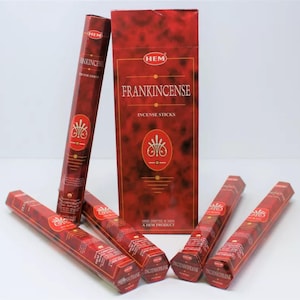 May include: A box of Hem Frankincense incense sticks with a red background and gold lettering. The box is surrounded by six individual incense sticks, each with a hexagonal cross-section.