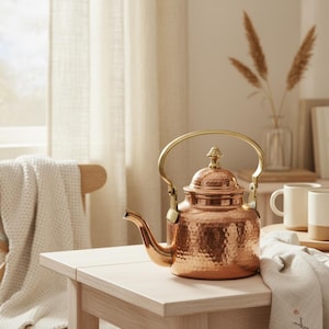 May include: A copper tea kettle with a hammered finish and a brass handle sits on a light wood table. The kettle has a decorative finial on the lid and a curved spout. The background includes a window, a chair, and a vase with dried plants.