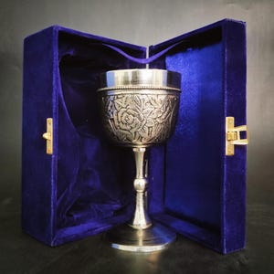 Könnte beinhalten: Ein silberner Kelch mit einem verzierten Blumenmuster befindet sich in einer blauen Samtbox mit einem goldenen Verschluss.