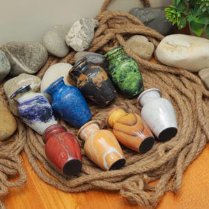 以下が含まれることがあります： さまざまな色と模様の小さな装飾用骨壺のコレクション。骨壺は研磨された素材で作られており、クラシックな花瓶の形をしています。色は青、緑、オレンジ、赤、白など。骨壺はロープの上に配置され、石に囲まれています。