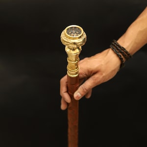 May include: A wooden walking stick with a brass compass top. The compass has a black face with white markings and a gold needle.