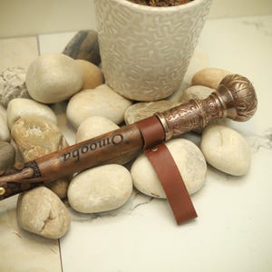 以下が含まれることがあります： 装飾的な金属製のハンドルと革製のストラップが付いた茶色の杖。「Omooba」の文字が木製のシャフトに刻まれています。ハンドルは詳細な模様で装飾されています。