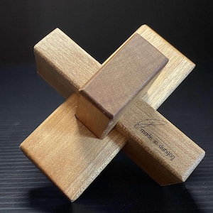 May include: A wooden puzzle in the shape of a cross with a small wooden block in the centre. The puzzle is made of light brown wood and has the text "made in dungog" on one side.