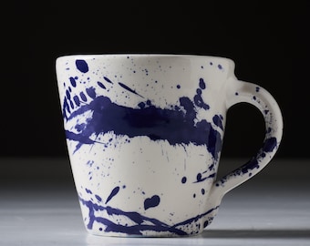 Puede incluir: Una taza de cerámica blanca con un diseño abstracto de salpicaduras azules y blancas. La taza tiene una forma redondeada y un asa.
