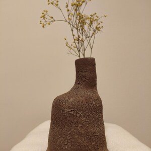 Könnte beinhalten: Eine strukturierte, braune Keramikvase mit einer einzigartigen, organischen Form. Die Vase enthält zarte, getrocknete gelbe Blumen, die eine natürliche und erdige Ästhetik erzeugen. Die Vase ist etwa 20 cm hoch, ideal für eine minimalistische Inneneinrichtung.
