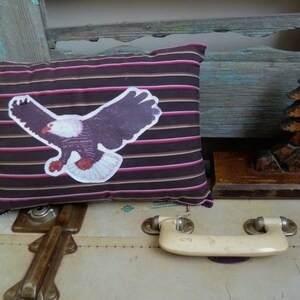 May include: A brown and pink striped pillow with a white eagle graphic. The pillow is sitting on top of a white suitcase with a brown handle.