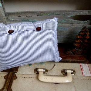 May include: A white and blue striped pillow with two brown buttons. The pillow is sitting on a vintage suitcase with a white leather exterior and a brass handle.
