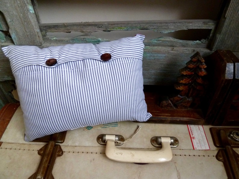 May include: A white and blue striped pillow with two brown buttons. The pillow is sitting on a vintage suitcase with a white leather exterior and a brass handle.