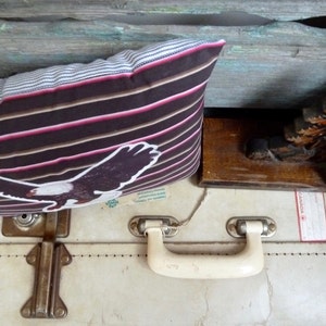 May include: A vintage suitcase with a brown and pink striped pillow with an eagle design on top. The suitcase has a white handle and two metal clasps. The suitcase has a label that says "Canada".