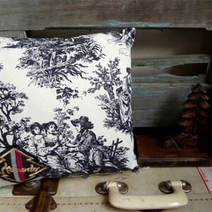 May include: A white cushion with a black and white toile pattern featuring a floral design and figures. The cushion has a black and red embroidered patch with the word "Remode" on it.