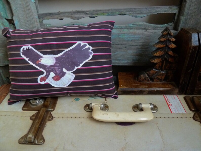 May include: A brown and pink striped pillow with a white eagle graphic. The pillow is sitting on top of a white suitcase with a brown handle.