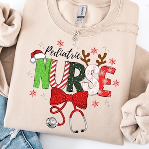 Puede incluir: Sudadera beige con la frase "Pediatric Nurse" en un diseño festivo. La palabra "NURSE" está decorada con un gorro de Papá Noel, cuernos de reno, copos de nieve y un lazo rojo con un estetoscopio. La sudadera está doblada.