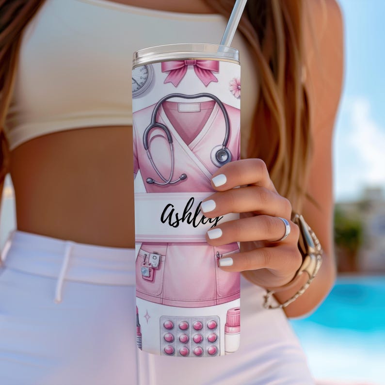 Puede incluir: Un vaso de acero inoxidable rosa y blanco con el nombre personalizado "Ashley" y un dise&ntilde;o que presenta un uniforme de enfermera, un estetoscopio, p&iacute;ldoras y otros suministros m&eacute;dicos.