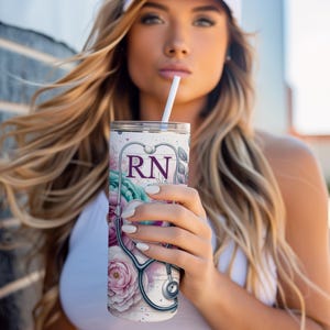 May include: A white stainless steel tumbler with a floral design and a stethoscope. The tumbler has the letters "RN" in purple on the front.