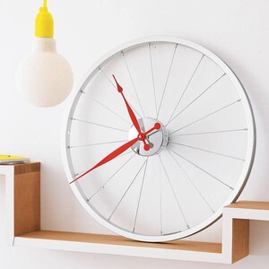 Può includere: Un orologio da parete bianco con un design a ruota di bicicletta. L'orologio ha una cornice argentata, sottili raggi e lancette rosse. L'orologio è montato su una parete bianca sopra una mensola di legno.