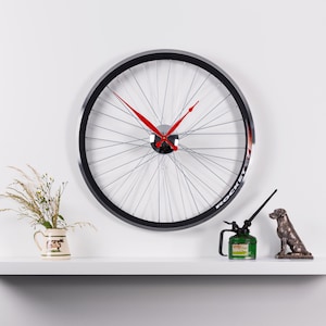 Peut inclure: Une horloge en forme de roue de vélo noire avec des aiguilles rouges. L'horloge est montée sur un mur blanc. Il y a un petit vase avec des fleurs et un petit bidon d'huile sur une étagère blanche sous l'horloge.