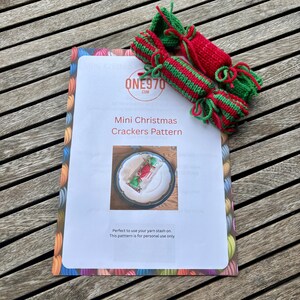 May include: A pattern for mini Christmas crackers with red and green knitted crackers on top. The pattern is on a white sheet with the text "ONE970.COM" and "Mini Christmas Crackers Pattern". The sheet is on a wooden surface.