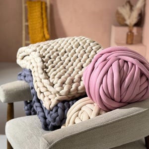 Op de afbeelding: Een stoel toont een stapel dikke gebreide dekens in crème, stoffig blauw en roze. De dikke, zachte textuur van de dekens wordt benadrukt. Een mosterdgele deken hangt op de achtergrond aan een houten ladder, wat wijst op een interieurthema.
