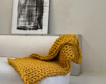 May include: A mustard yellow chunky knit blanket draped over a white, curved sofa. The blanket's thick weave is visible, creating a textured appearance. An abstract black and white painting hangs on the wall behind the sofa, adding a modern touch.