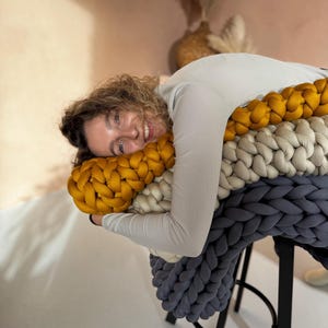 May include: A woman is lying on a large, chunky knit pillow. The pillow is made of three colors: mustard yellow, cream, and dark gray. The pillow is on a black stool.
