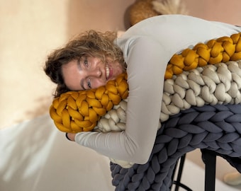 May include: A woman is lying on a large, chunky knit cushion. The cushion is made of three colours: mustard yellow, cream, and dark grey. The cushion is on a black stool.