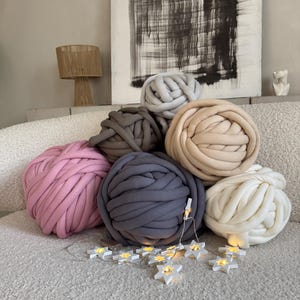 Peut inclure: Plusieurs grosses boules de fil à tricoter épais dans des tons de rose, de gris et de crème. Une guirlande de lumières en forme d'étoiles est drapée au premier plan. Les boules de fil sont disposées sur une surface claire et texturée.