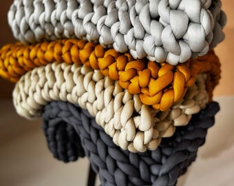 May include: A stack of four chunky knit blankets in various colors: gray, mustard yellow, cream, and charcoal. The blankets are made of thick, soft yarn and are arranged on top of each other. The blankets are displayed on a black stool.