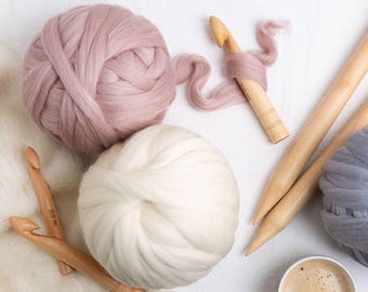 May include: A collection of craft supplies including large balls of yarn in pink, white, and gray, wooden crochet hooks, and knitting needles. A cup of coffee sits nearby. The yarn appears to be a chunky merino wool.