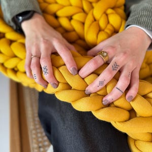 Könnte beinhalten: Nahaufnahme einer senfgelben, grob gestrickten Decke. Die Decke ist dick und geflochten, mit einer weichen Textur. Die Hände einer Person liegen auf der Decke, mit Tattoos und Ringen sichtbar. Die Person trägt eine schwarze Jeans und eine grüne Jacke.