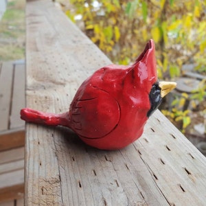 May include: A red cardinal bird figurine made of clay with a yellow beak. The figurine is sitting on a wooden surface.