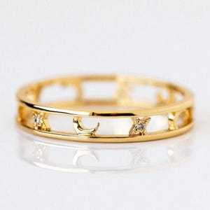 Könnte beinhalten: Ein goldfarbener Ring mit einem Himmelsdesign. Der Ring hat ausgeschnittene Abschnitte mit Stern-, Halbmond- und Sternform. Kleine klare Edelsteine sind in die Sternmuster eingesetzt. Der Ring wird auf einer weißen Oberfläche präsentiert.